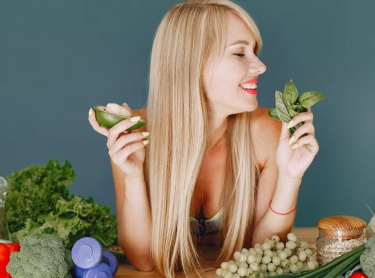 A smiling woman holding avocado and basil, surrounded by fresh vegetables and fruits for healthy eating.