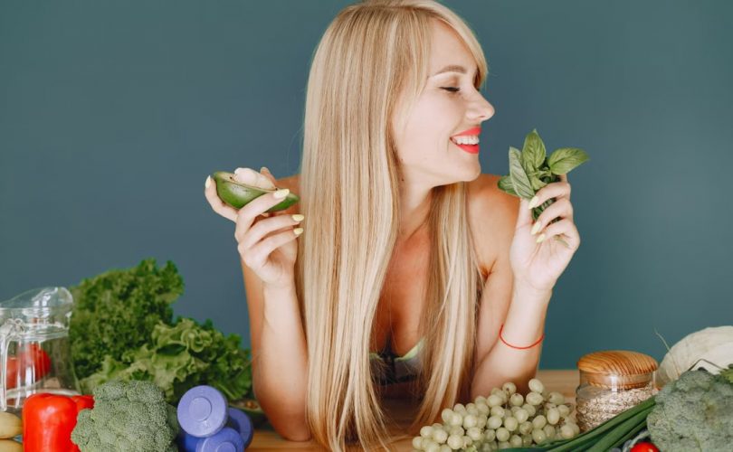 A smiling woman holding avocado and basil, surrounded by fresh vegetables and fruits for healthy eating.
